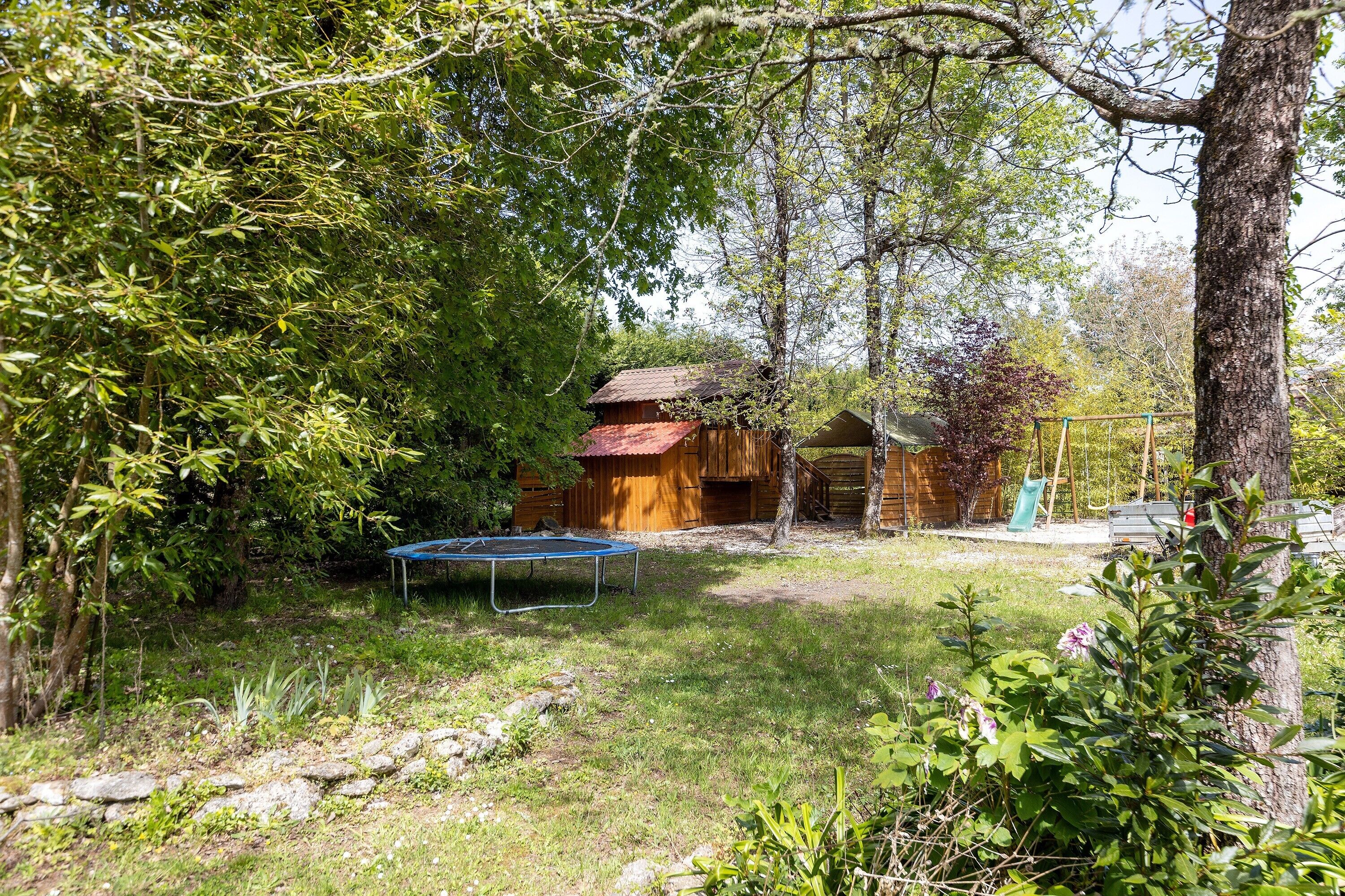 A rustic wooden cabin surrounded by dense trees and a trampoline in a grassy yard.
