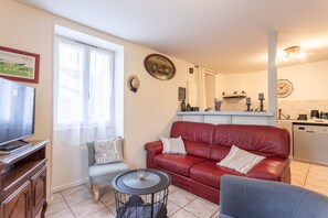 A living room with a red leather sofa, a grey armchair, a small round table, a television, and a kitchen area with a white countertop and appliances. - Apartment 'Etxexuria' with Private Terrace, Wi-Fi and Air Conditioning (Cambo les Bains)