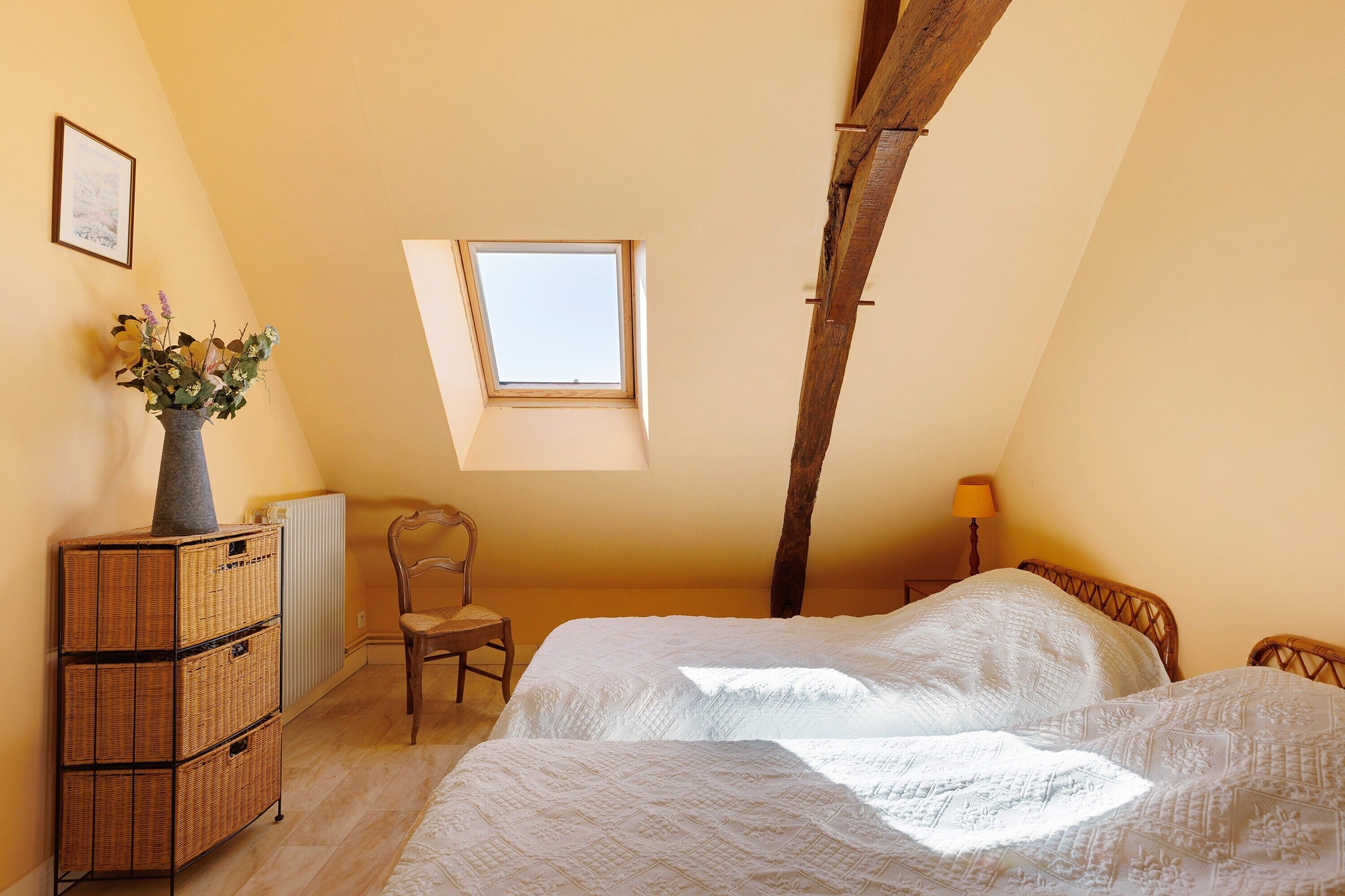 A bedroom with a sloped ceiling, a skylight, a bed with white bedding, a wooden dresser, and a chair.