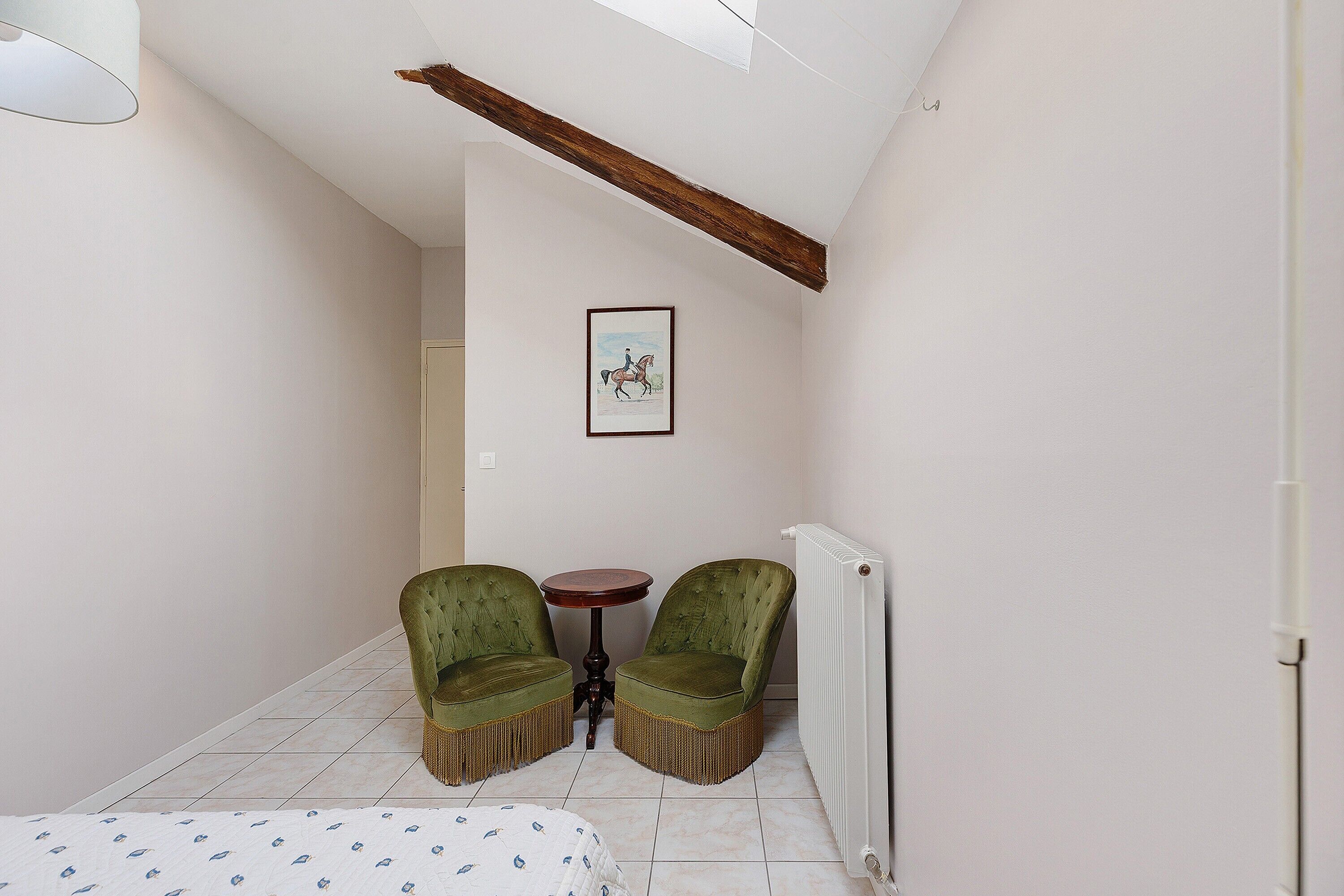 A room with a bed, two green chairs, a small table, a framed picture on the wall, and a radiator.