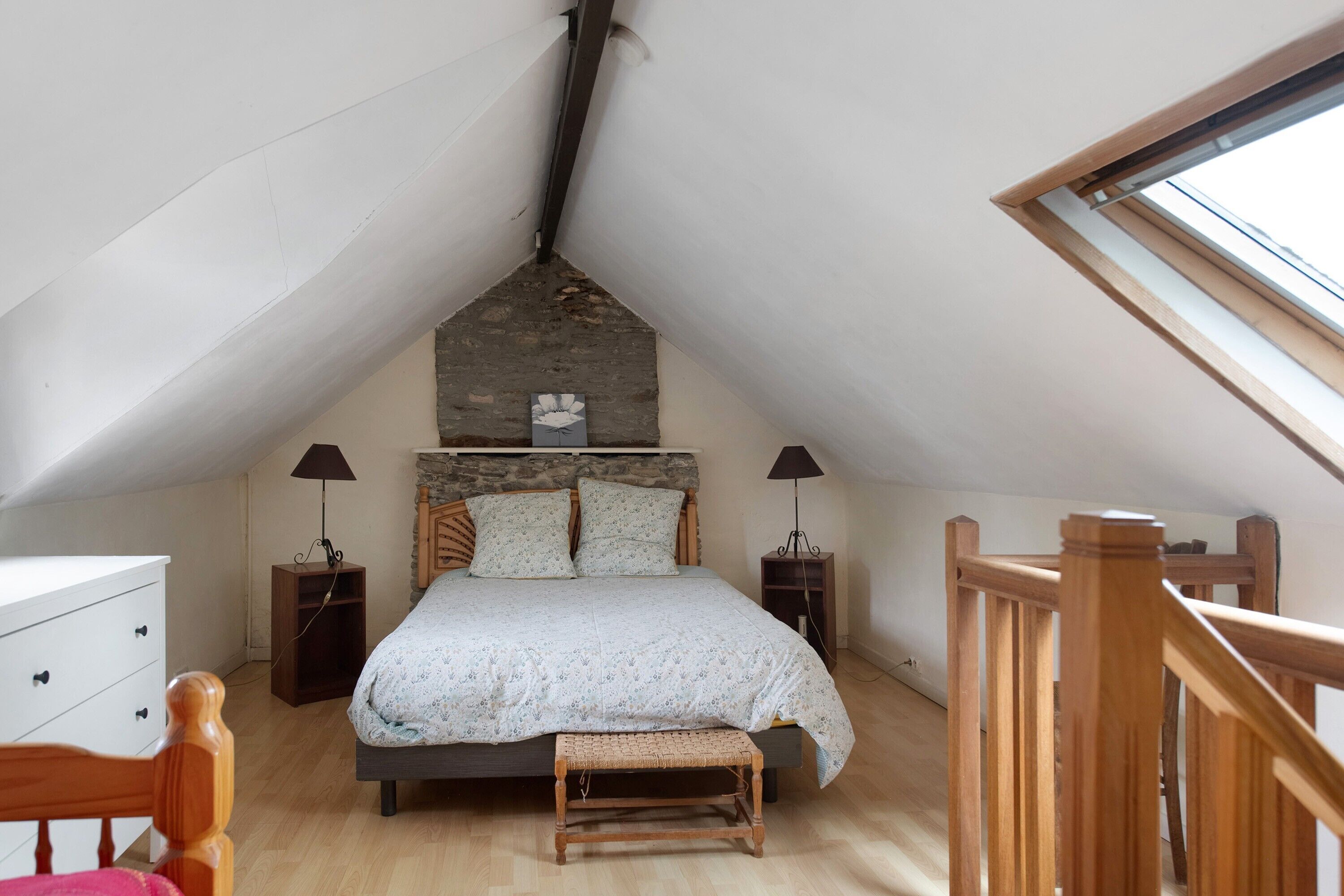Un dormitorio con techo inclinado, una cama con cabecera, dos mesitas de noche, un banco y una ventana.