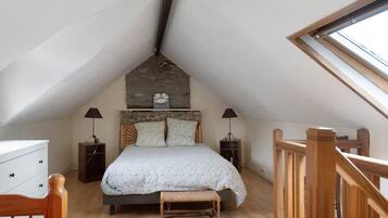 Un dormitorio con techo inclinado, una cama con cabecera, dos mesitas de noche, un banco y una ventana.