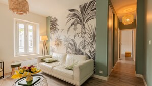 Een woonkamer met een bank, een bijzettafeltje met bloemen, een lamp en een muur met een tropische muurschildering.