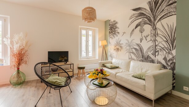 Een woonkamer met een bank, een fauteuil, een salontafel, een televisie en een muur met een tropisch schilderwerk.