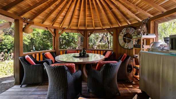 Un espace extérieur abrité, aménagé avec des chaises en osier et une table ronde, entouré de poutres en bois et offrant une vue sur la verdure.