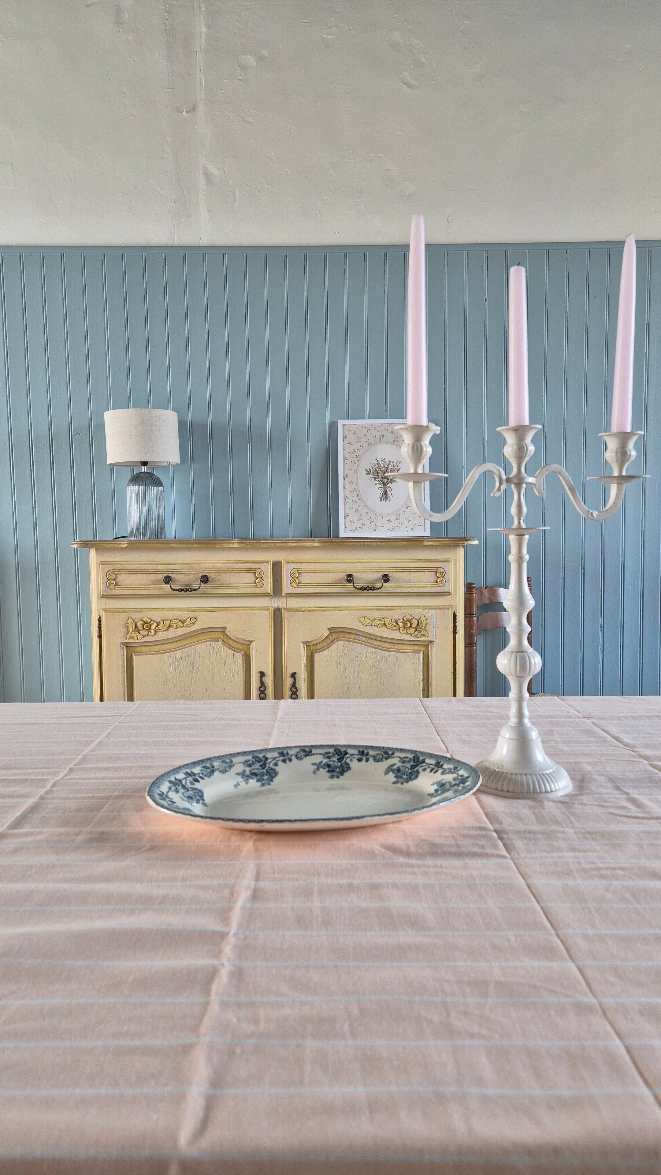A table with a plate, a candelabra with candles, a lamp, and a dresser with a picture frame against a blue wall.