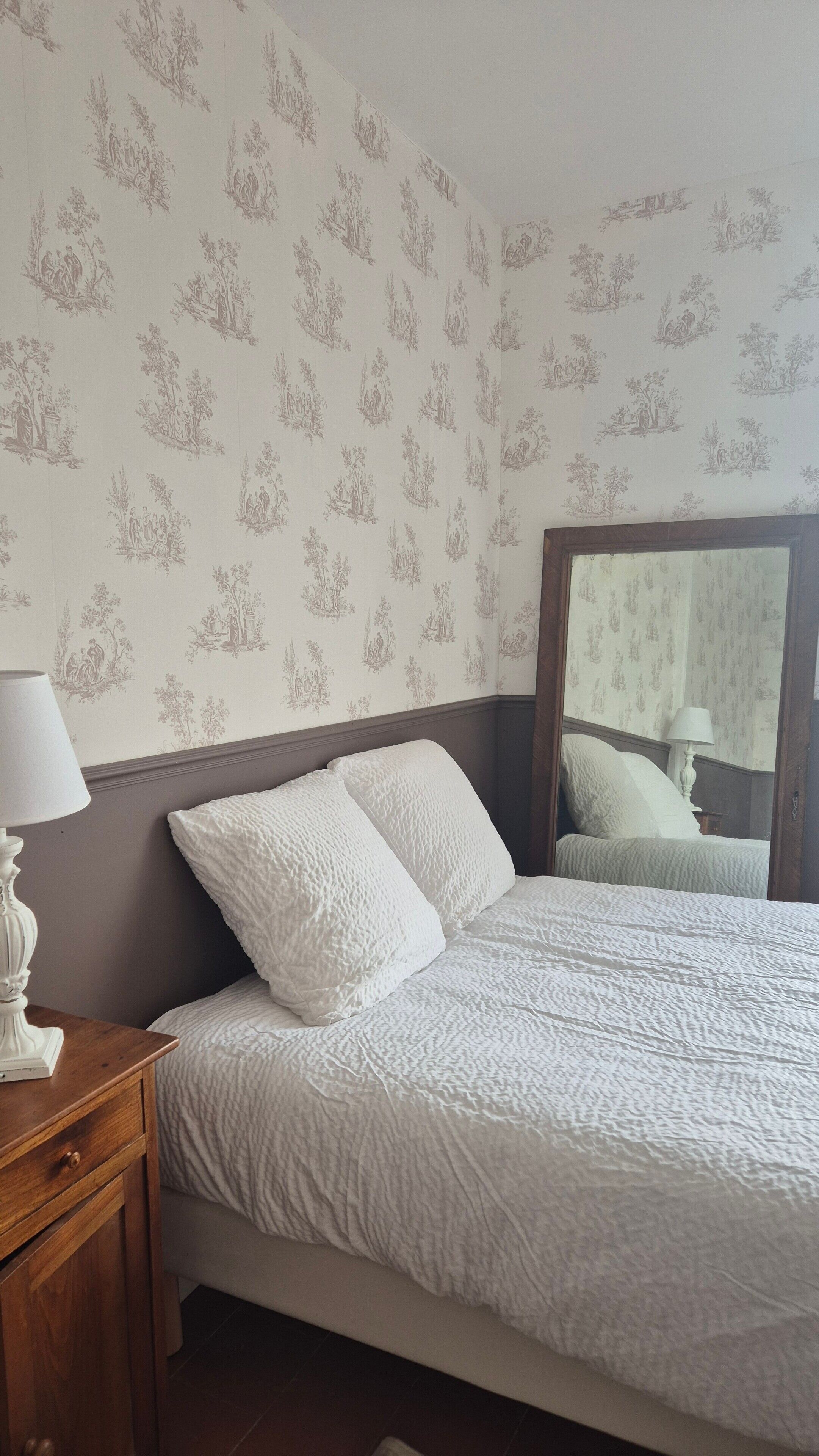 A bedroom with a bed, a nightstand, a mirror, and wallpaper with a repeating pattern.