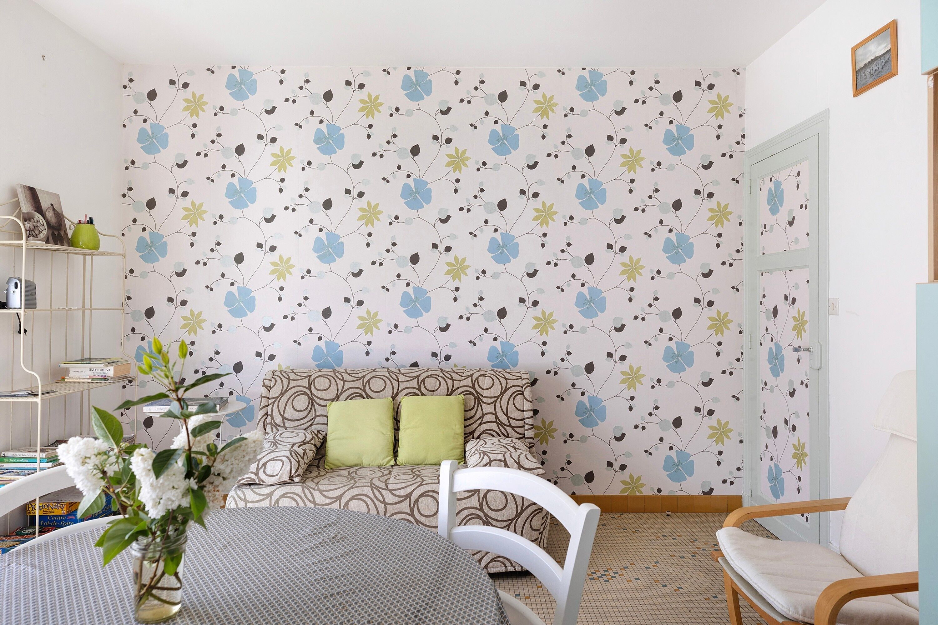A room with a patterned wallpaper, a sofa with cushions, a table with a vase, and a chair.