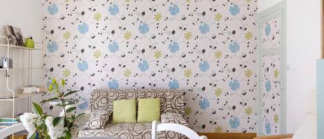 A room with a patterned wallpaper, a sofa with cushions, a table with a vase, and a chair.
