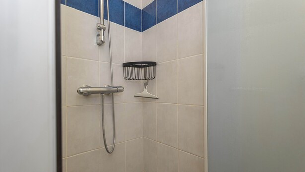 A shower with a handheld showerhead and a wall-mounted showerhead, a black shower caddy, and a corner tile design with blue accents. - Maison Goéland – A Relaxing Getaway Between Land and Sea, Shared Pool & Balcony (Le Barcarès)