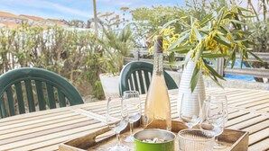 A wooden table with a bottle, glasses, and a bowl, set outdoors with chairs and plants in the background. - Maison Goéland – A Relaxing Getaway Between Land and Sea, Shared Pool & Balcony (Le Barcarès)