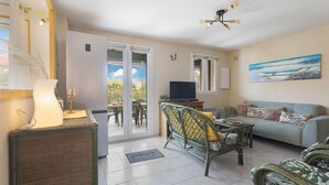 A living room with a sofa, a television, wicker furniture, and a painting on the wall. - Maison Goéland – A Relaxing Getaway Between Land and Sea, Shared Pool & Balcony (Le Barcarès)
