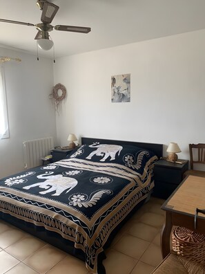 Une chambre avec un lit, un ventilateur de plafond, une œuvre d’art murale, une décoration murale tissée et une petite table avec une lampe.