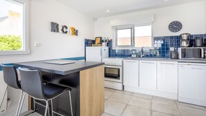 A modern kitchen with a central island, white cabinets, a blue backsplash, and a clock on the wall. - Holiday Home 'L'escale bleue' with Shared Terrace, Private Garden & Wi-Fi (Plomeur)
