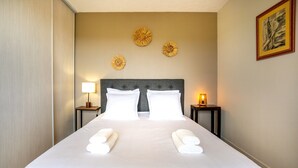 A neatly made bed with white linens and towels, a grey upholstered headboard, and decorative wall art. - Holiday Home 'L'escale bleue' with Shared Terrace, Private Garden & Wi-Fi (Plomeur)