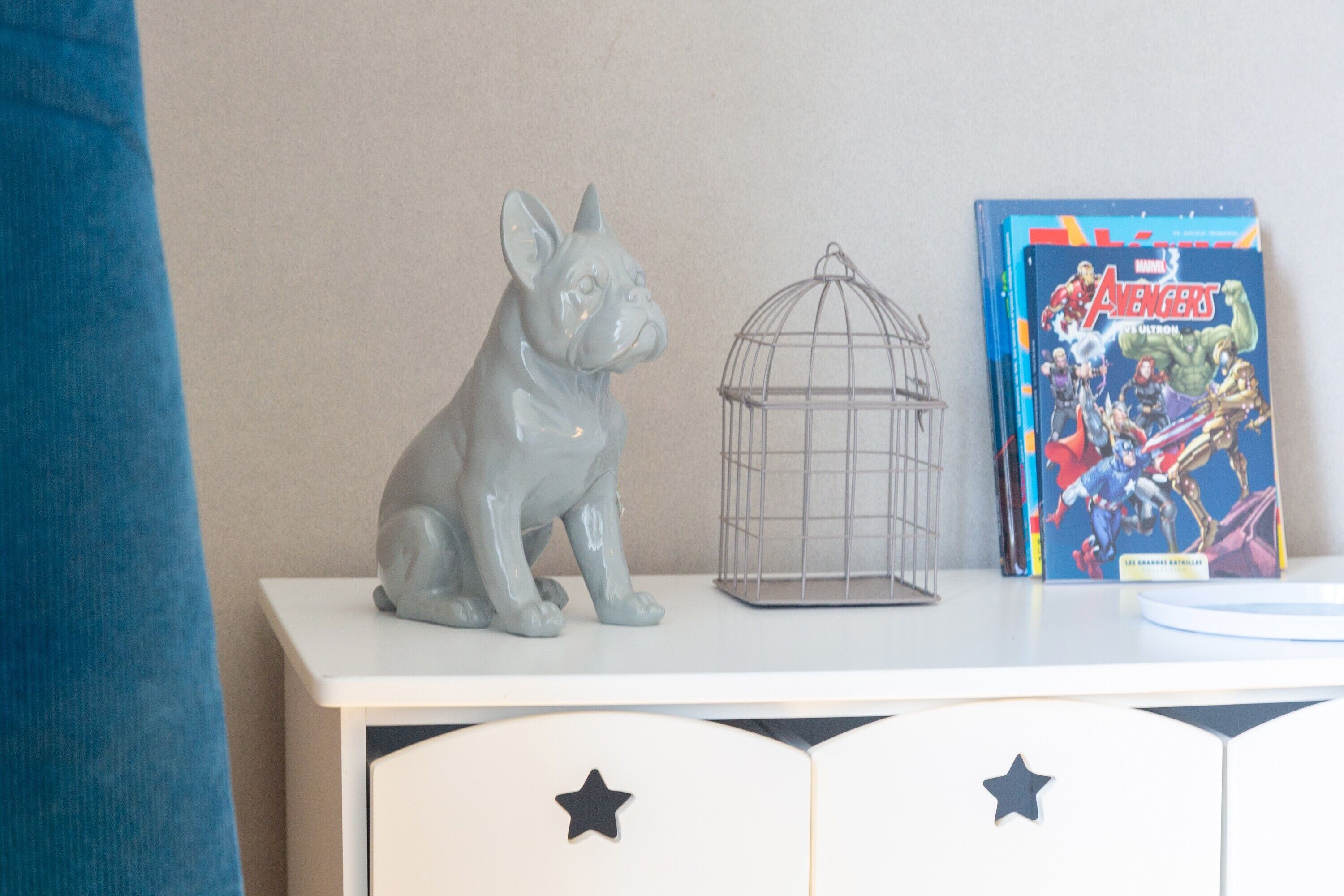 A white cabinet with star decorations, a grey dog figurine, a wire birdcage, and a Marvel Avengers comic book.