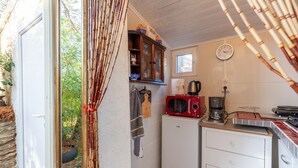 Une cuisine compacte équipée d’un réfrigérateur, d’un four à micro-ondes et d’une machine à café. On aperçoit une fenêtre avec des rideaux, et une horloge au mur indique l’heure.