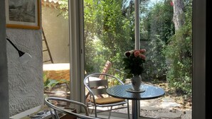 Une pièce avec une porte vitrée donnant sur un jardin, une table ronde entourée de chaises, un tableau encadré accroché au mur et un banc en bois.
