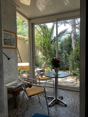 A room with a glass door leading to a garden, a round table with chairs, a framed picture on the wall, and a wooden bench.