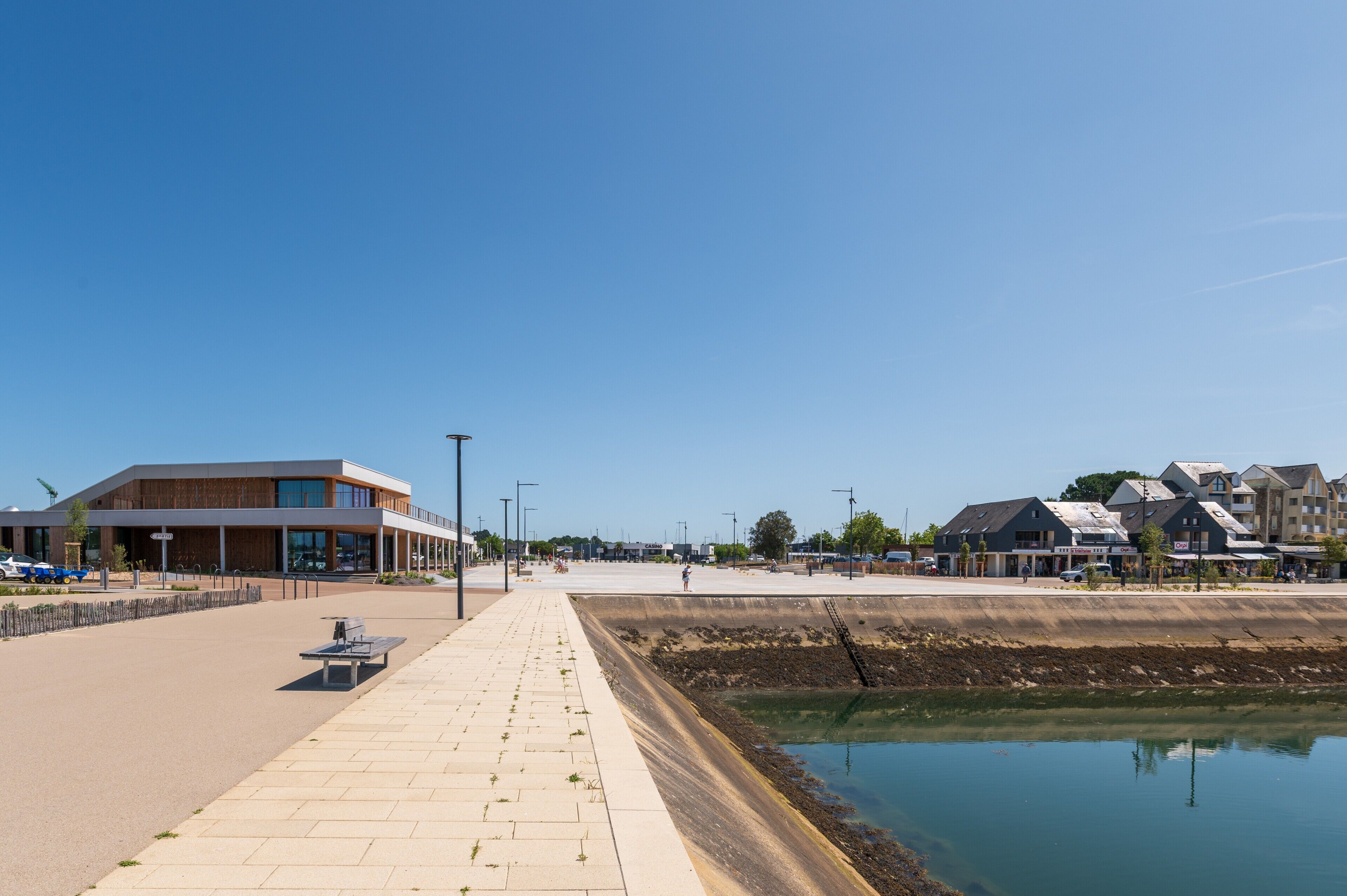 Une promenade en bord de mer avec un banc, un bâtiment doté de grandes fenêtres et une étendue d’eau calme.