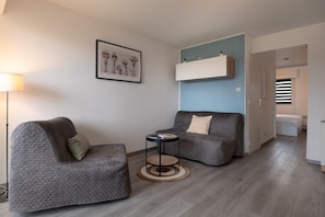 A modern living room with a grey sofa, a small round table, a floor lamp, and a wall-mounted shelf with decorative items. - Le Sailing Apartment: Port View, Near Beach & Thalasso, Private Parking (Arzon)