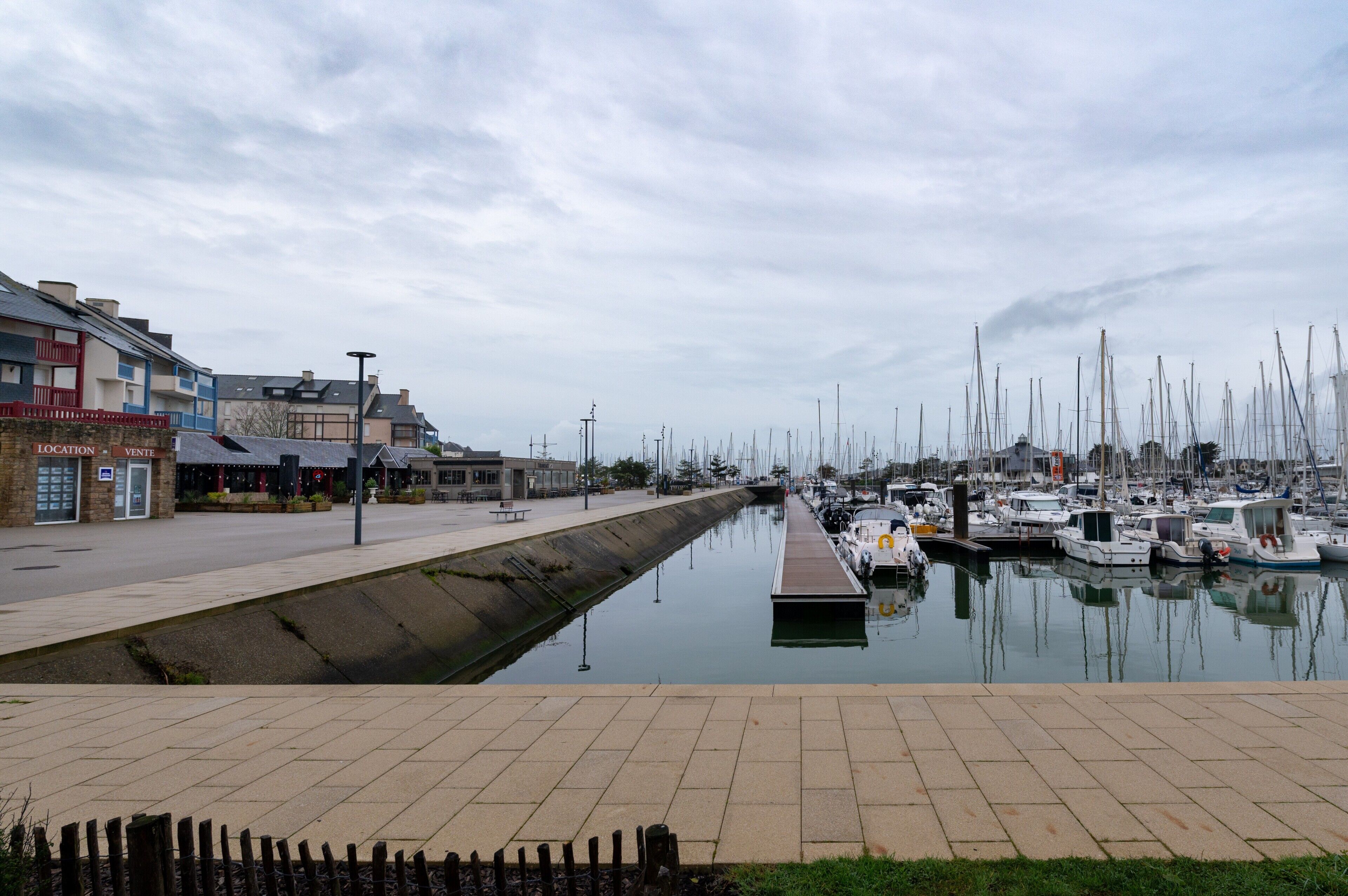 A marina with boats docked, a canal, and a row of buildings with a sign that reads 'LOCATION VENT'.