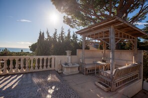 Ein hölzerner Pavillon mit einer Bank und einem Tisch, eine Steinfountain und eine geflieste Terrasse mit Meerblick.