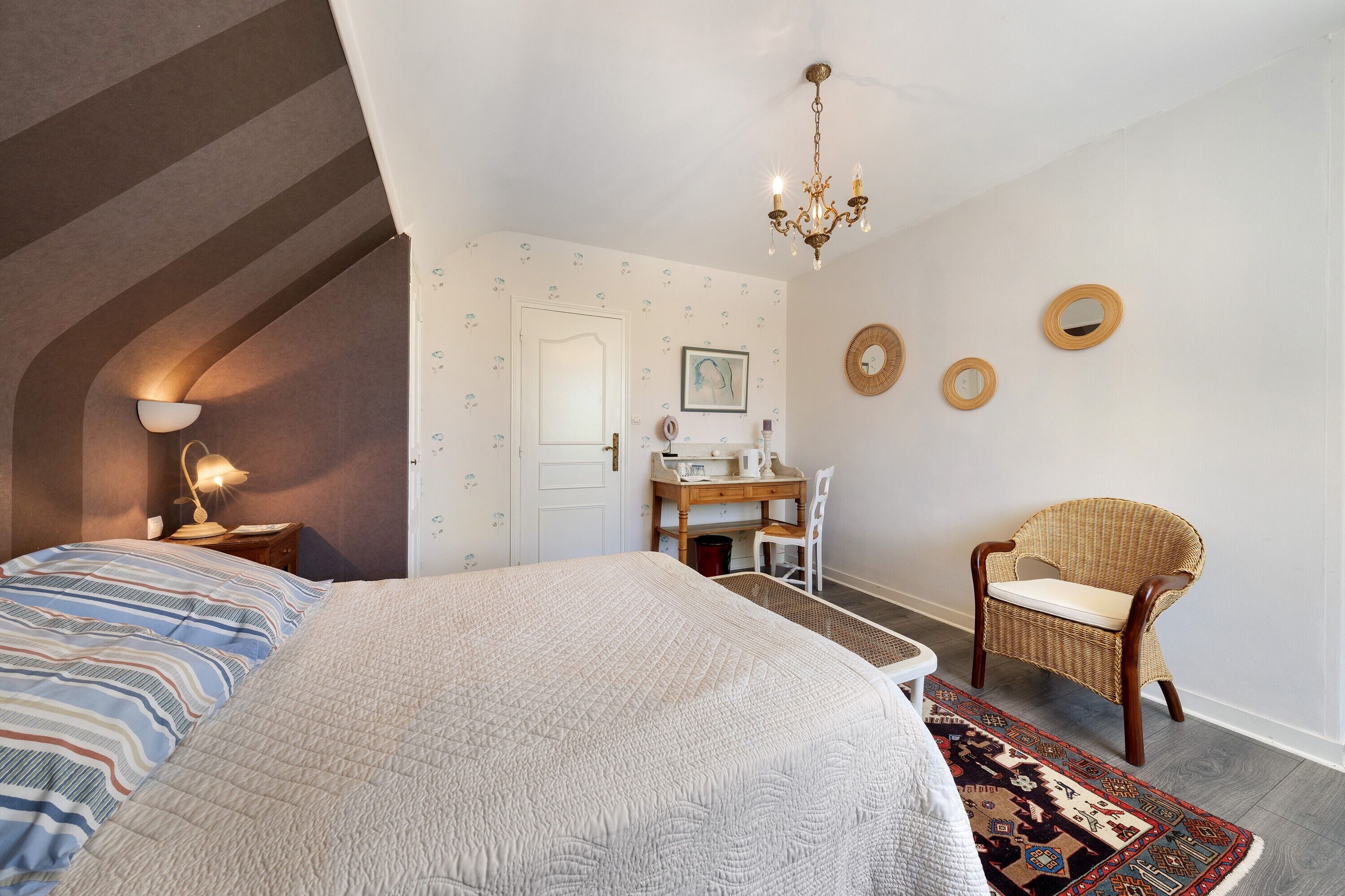 A bedroom with a bed, a desk, a chair, a chandelier, and a patterned rug.