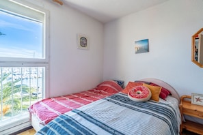 Ein Schlafzimmer mit großem Fenster, einem Bett mit gemusterten Betttextilien, einem kleinen Nachttisch und einem Wandbild.