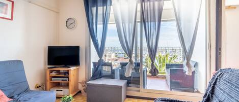 A living room with a sofa, TV, ottoman, and a balcony with curtains.