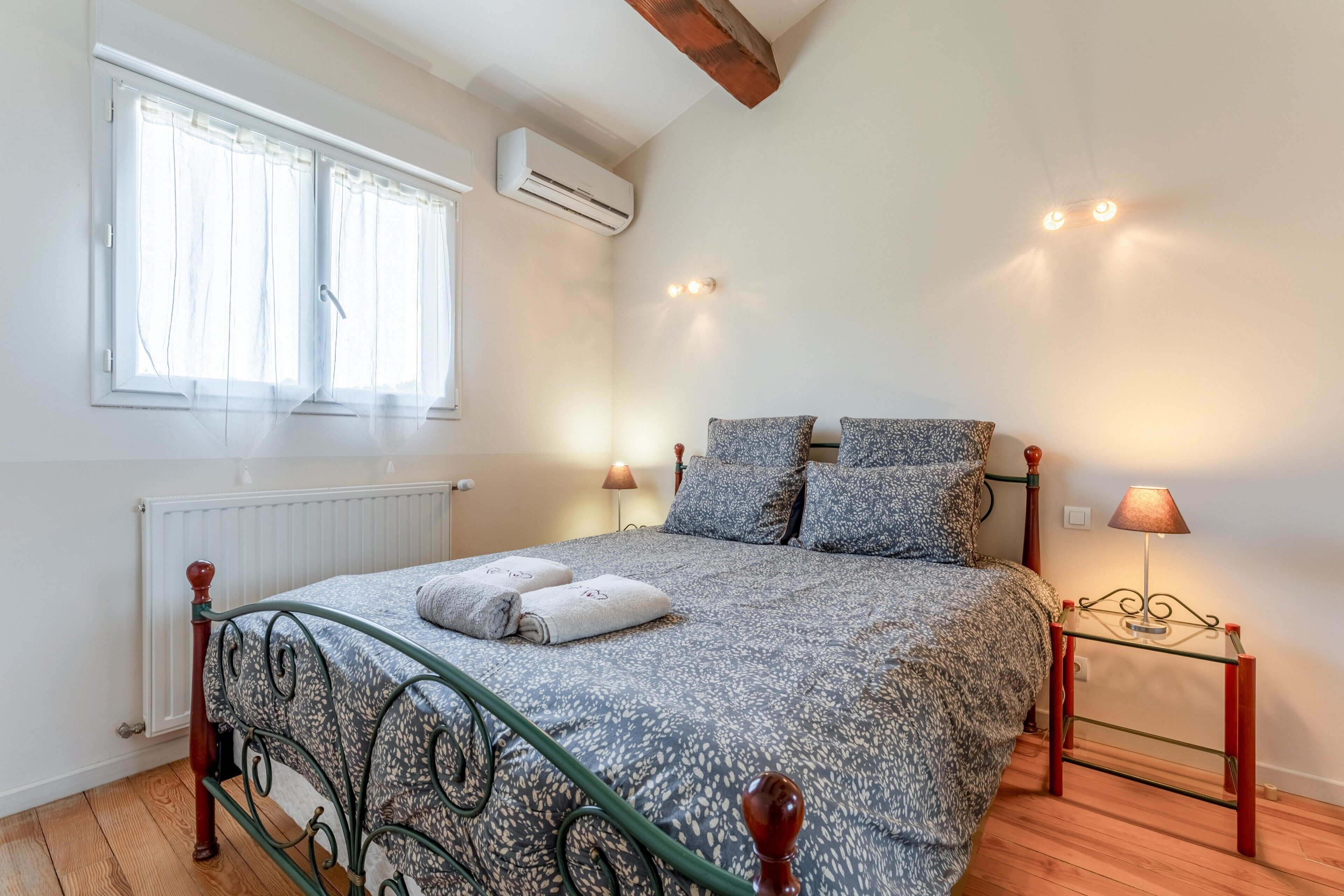 A bedroom with a bed, two bedside lamps, a window with curtains, an air conditioning unit, and a wooden floor.