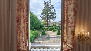 Una habitación con vistas a través de una ventana con cortinas, un radiador, un sillón de mimbre y una mesa de vidrio.