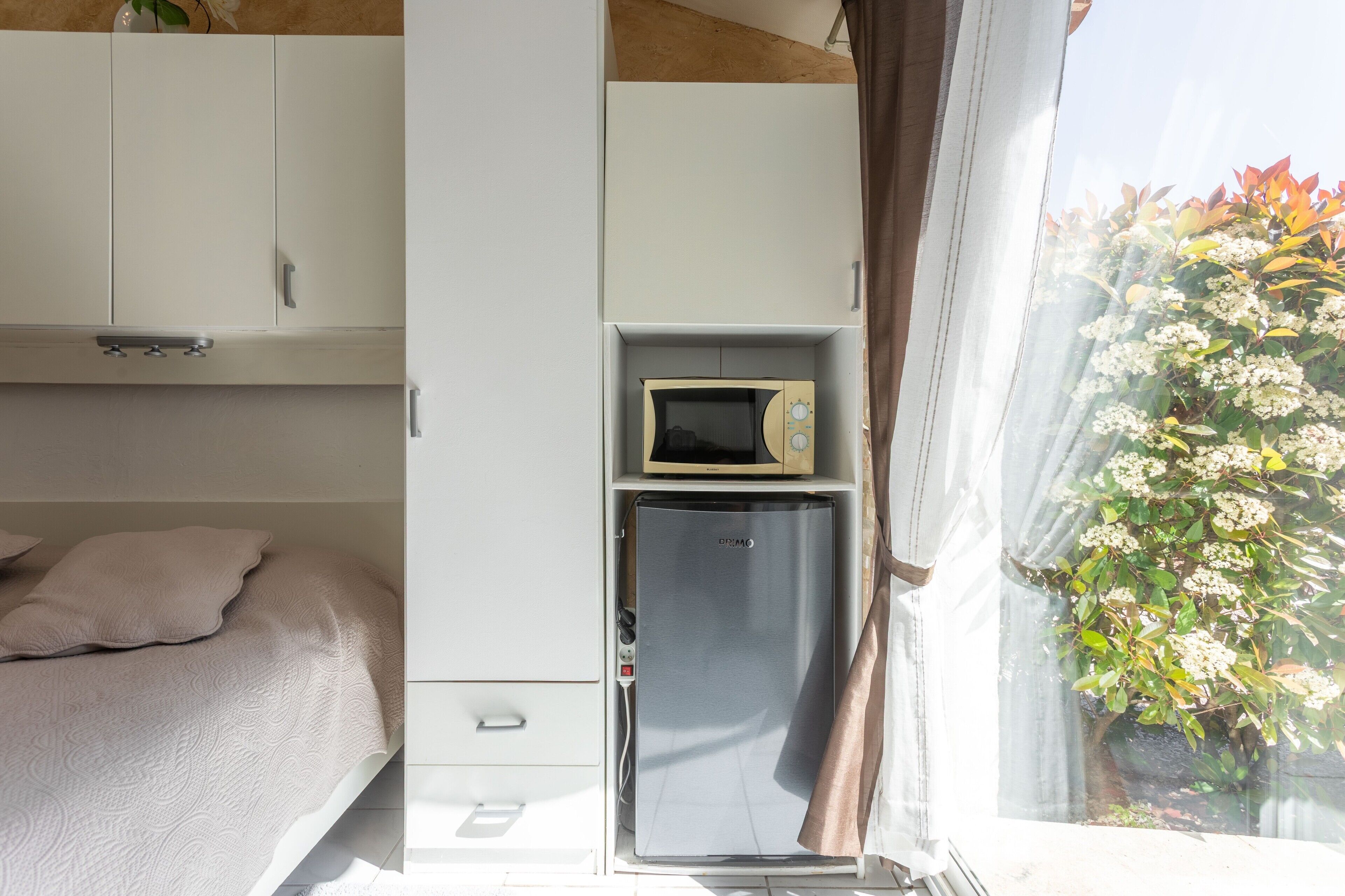 Une chambre à coucher comprenant un lit, une armoire blanche, un petit réfrigérateur et une fenêtre donnant sur un paysage verdoyant.
