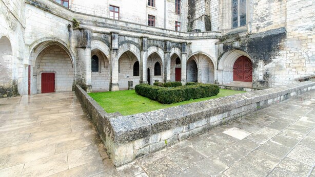 Un cortile con archi in pietra, un cespuglio al centro e una porta rossa.