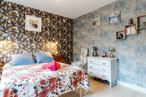 Un dormitori amb un paper pintat de motius florals, un llit amb llençols florits, un calaixera blanc i prestatges fixats a la paret amb objectes decoratius.