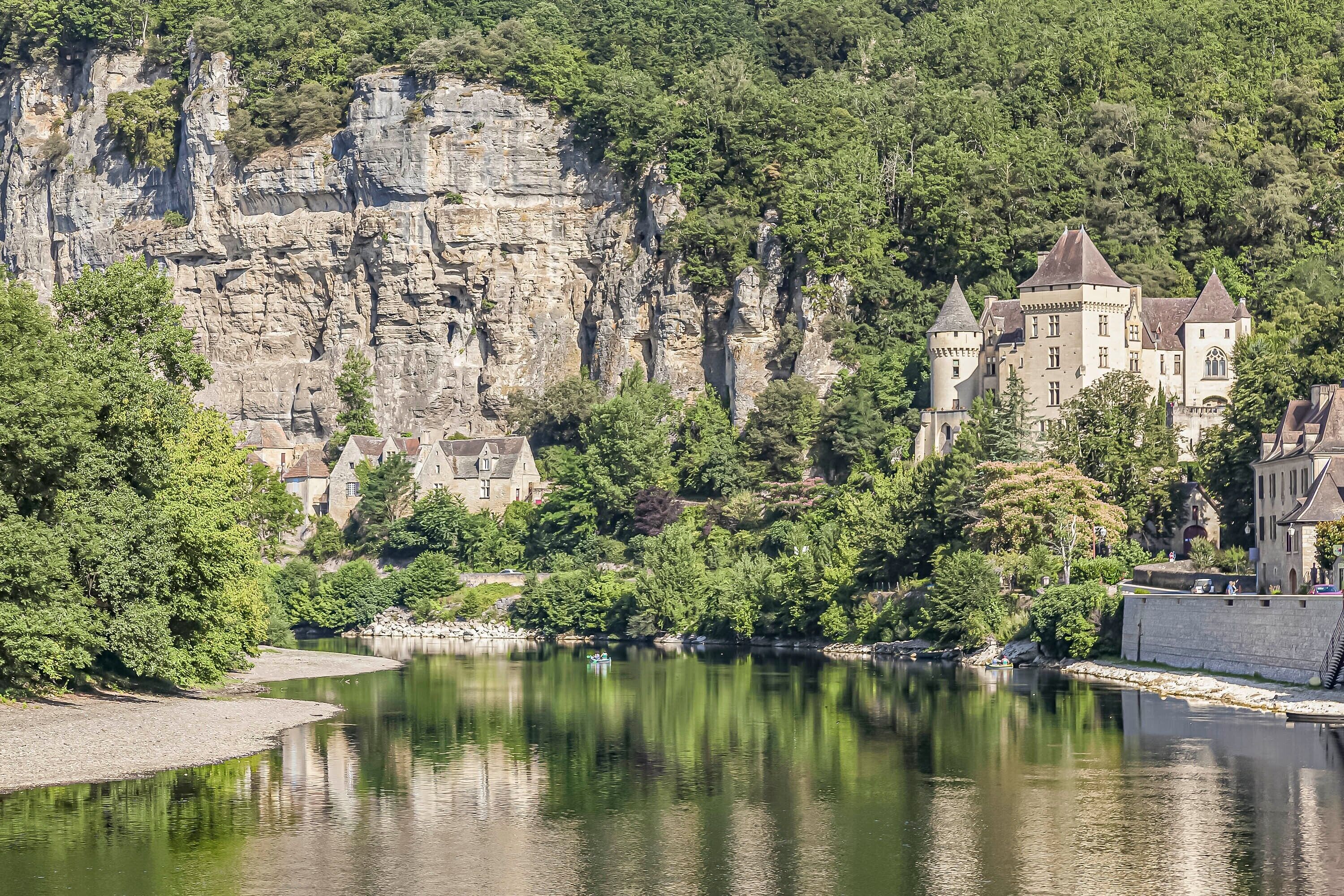 A castle-like building with multiple towers, situated by a calm river, surrounded by dense greenery and rocky cliffs.