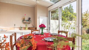 Et spiseområde med et bord dækket til fire personer, en vase med blomster og udsigt til haven gennem store vinduer.