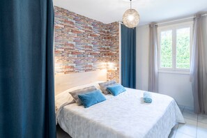 Une chambre avec un mur décoré d’un motif en briques, un lit avec des coussins bleus, un luminaire suspendu et une fenêtre avec des rideaux.