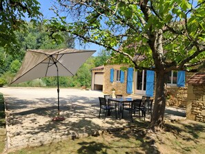 Un espace extérieur aménagé pour les repas, avec une table et des chaises, un grand parasol et un bâtiment en pierre doté de volets bleus.
