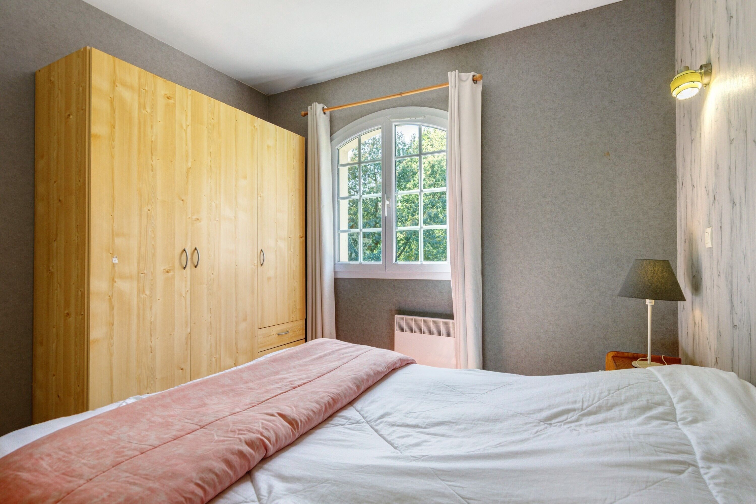 Ein Schlafzimmer mit Bett, einem Holzschrank, einem Fenster mit Vorhängen und einer Nachttischlampe.