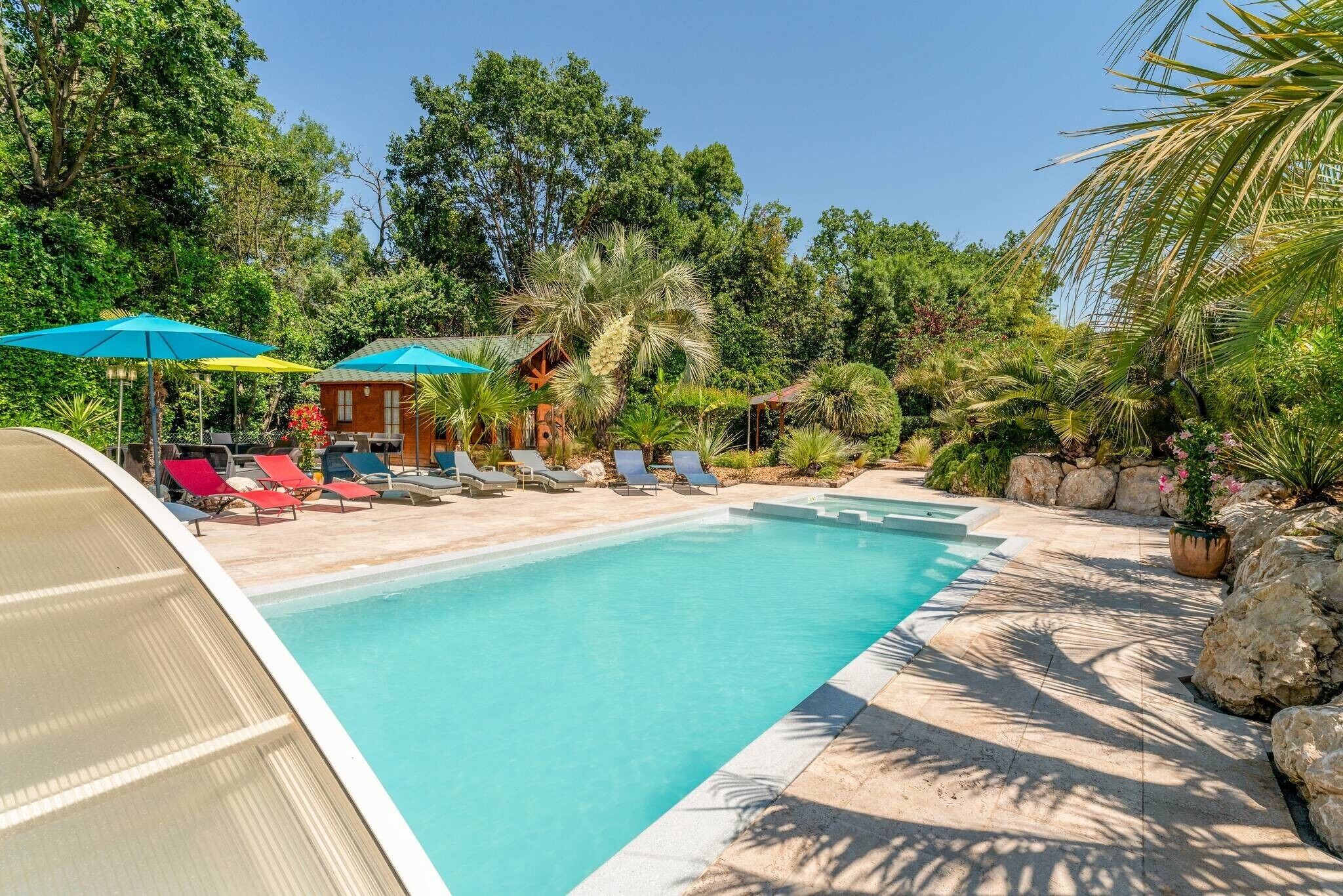 A swimming pool with lounge chairs and umbrellas, surrounded by greenery and rocks.