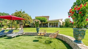 Une pelouse bien entretenue, agrémentée de chaises longues et d’un parasol rouge, un mur de pierre et, au fond, une maison avec un toit en tuiles.