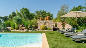 Une piscine entourée de verdure et de constructions en pierre, avec des chaises longues et un parasol.