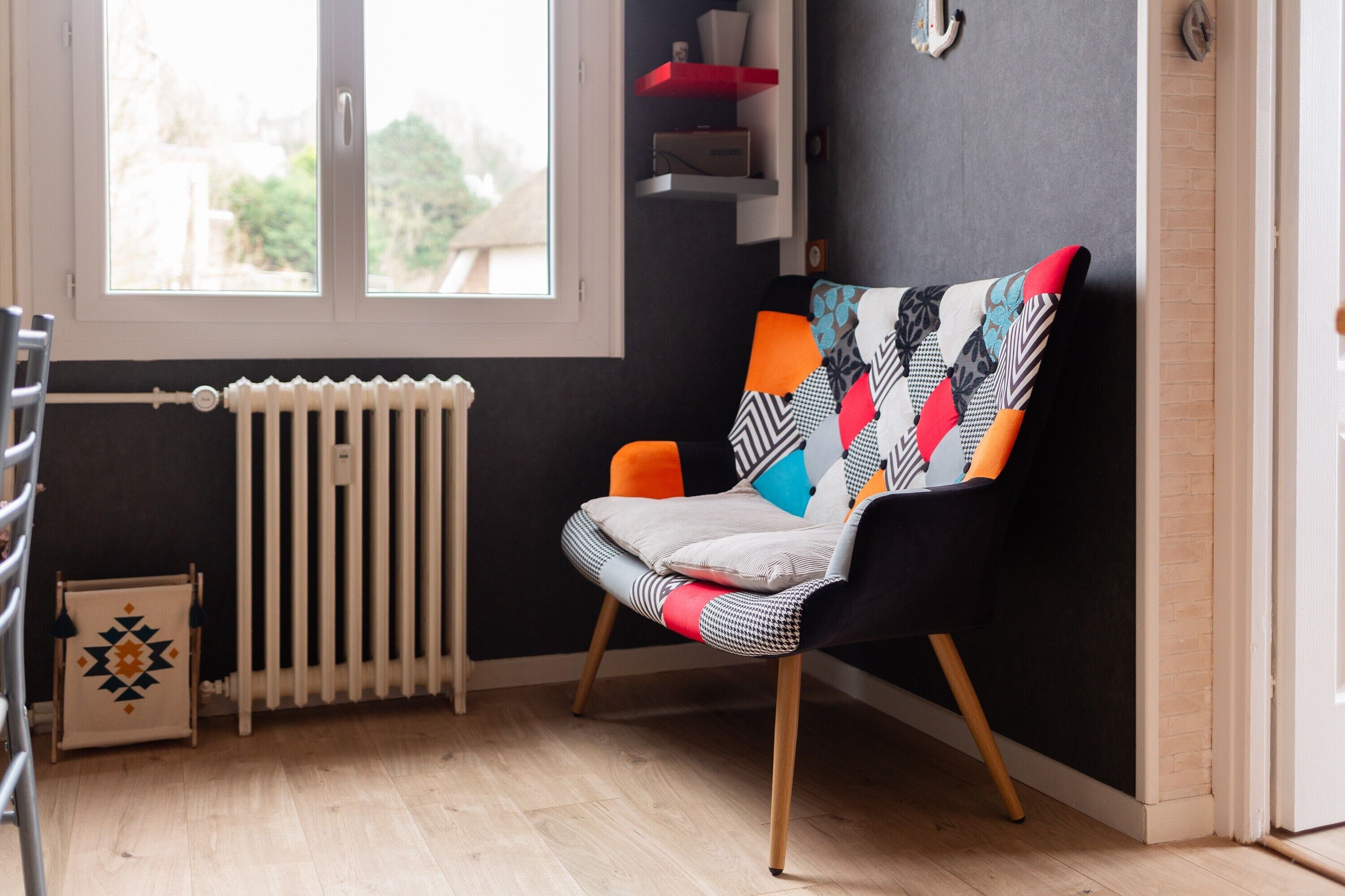 A modern room with a black wall, a window, a radiator, a chair with a colorful cushion, and a small shelf with decorative items.