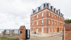 Een meerlaags bakstenen gebouw met een hekpoort, een klein buitengebied met treden, en uitzicht op zee.