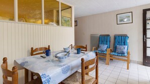 Uma sala de jantar com uma mesa posta para dois, cadeiras de madeira, toalha de mesa estampada em azul e branco, e uma janela com cortinas.