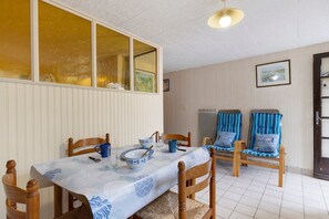 Ein Essbereich mit einem Tisch für zwei Personen, Holzstühlen, einer blau-weißen Tischdecke und einem Fenster mit Vorhängen.