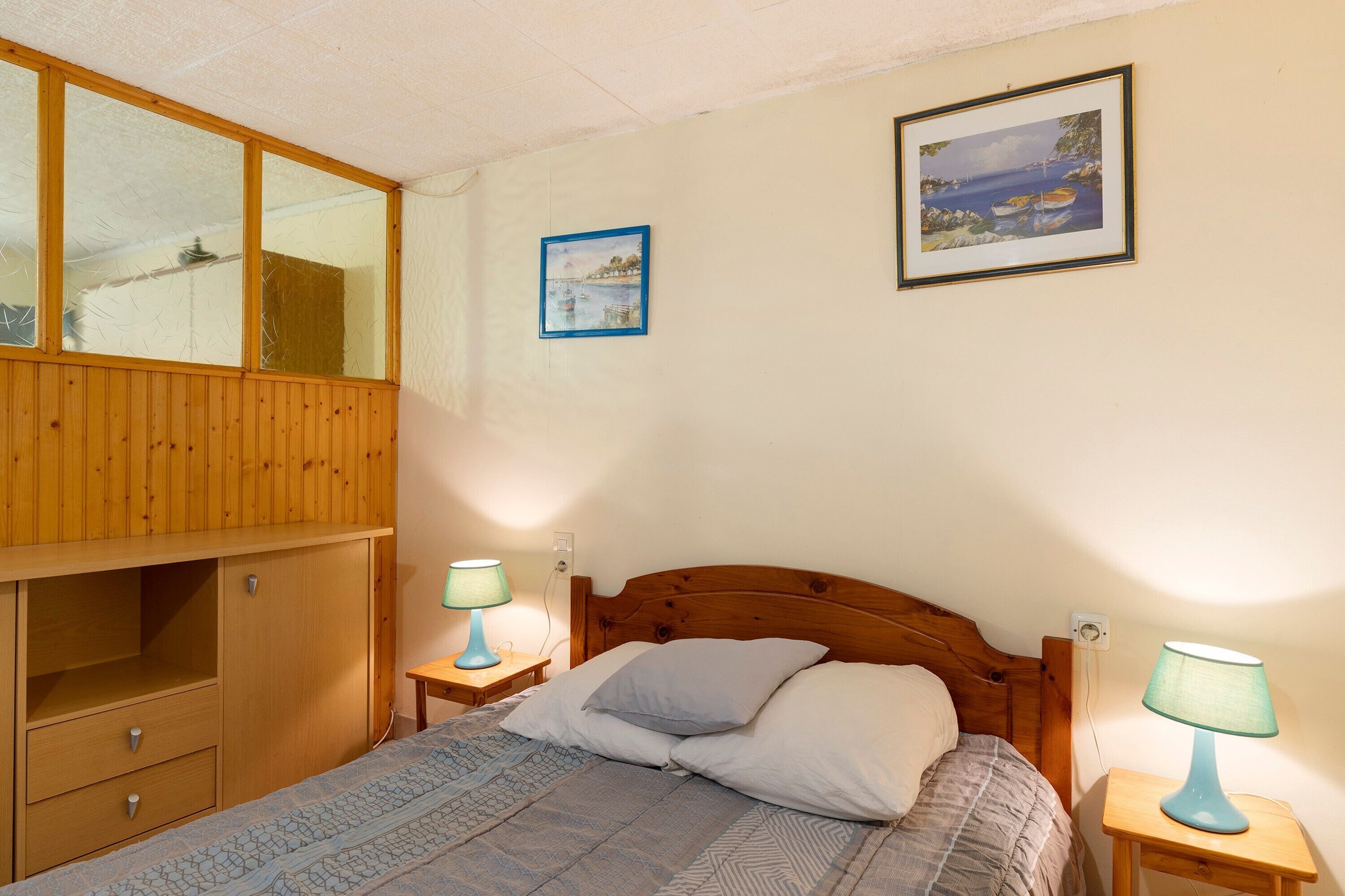 A bedroom with a wooden headboard, a bed with a patterned bedspread, two bedside tables with lamps, a wooden wardrobe, and two framed pictures on the wall.