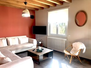 A living room with a sofa, a coffee table, a chair, a TV, a window, and a radiator. - Gîte "Fleur de Miel" – Renovated House in the Heart of the Lakes Region (Saffloz)