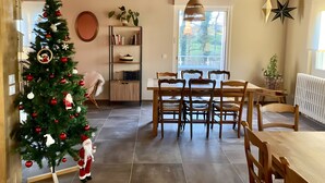 Un coin repas avec une table et des chaises en bois, un sapin de Noël, une bibliothèque et une fenêtre donnant sur la verdure.
